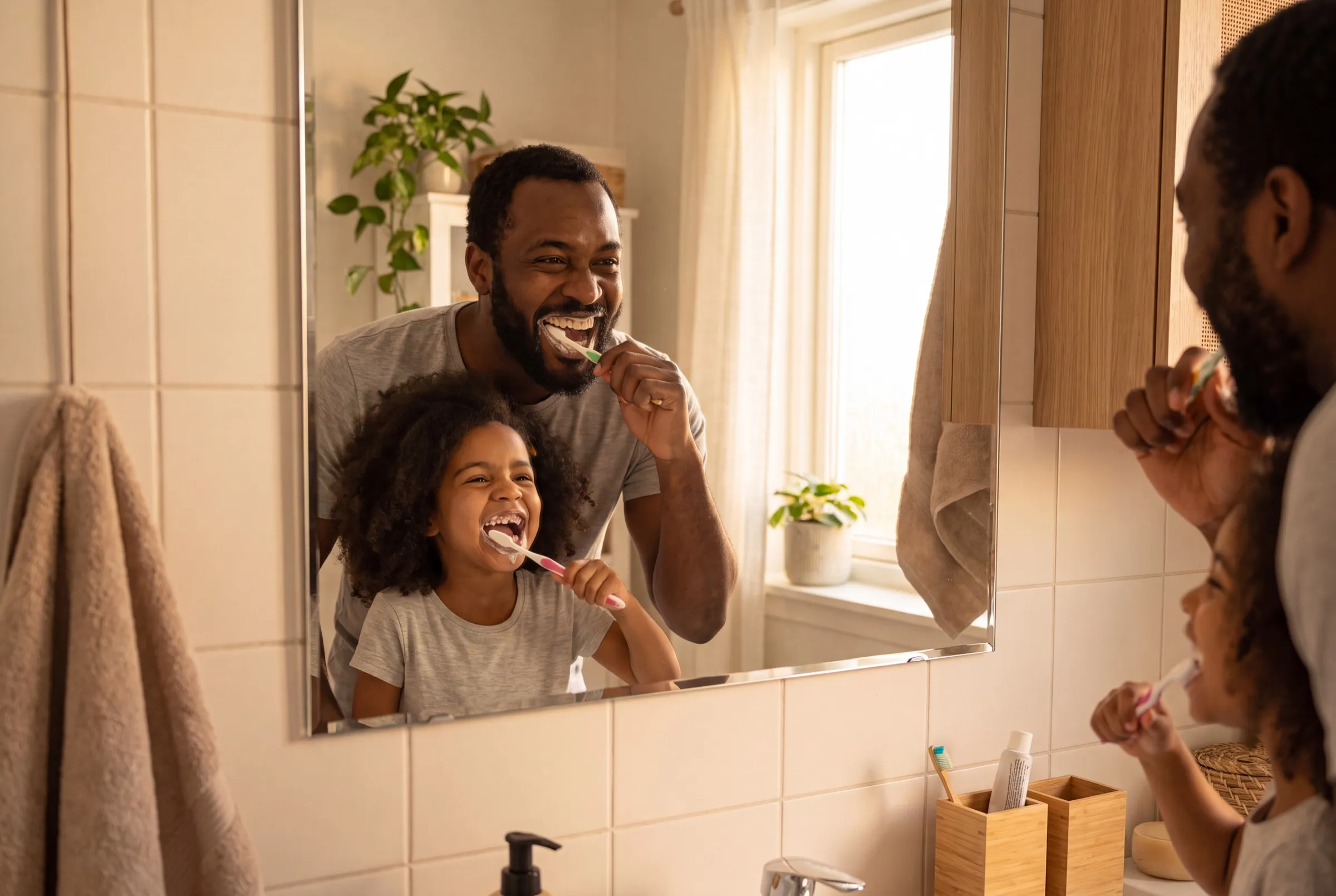 A father and his young daughter brushing their teeth side by side at a bathroom mirror in morning light