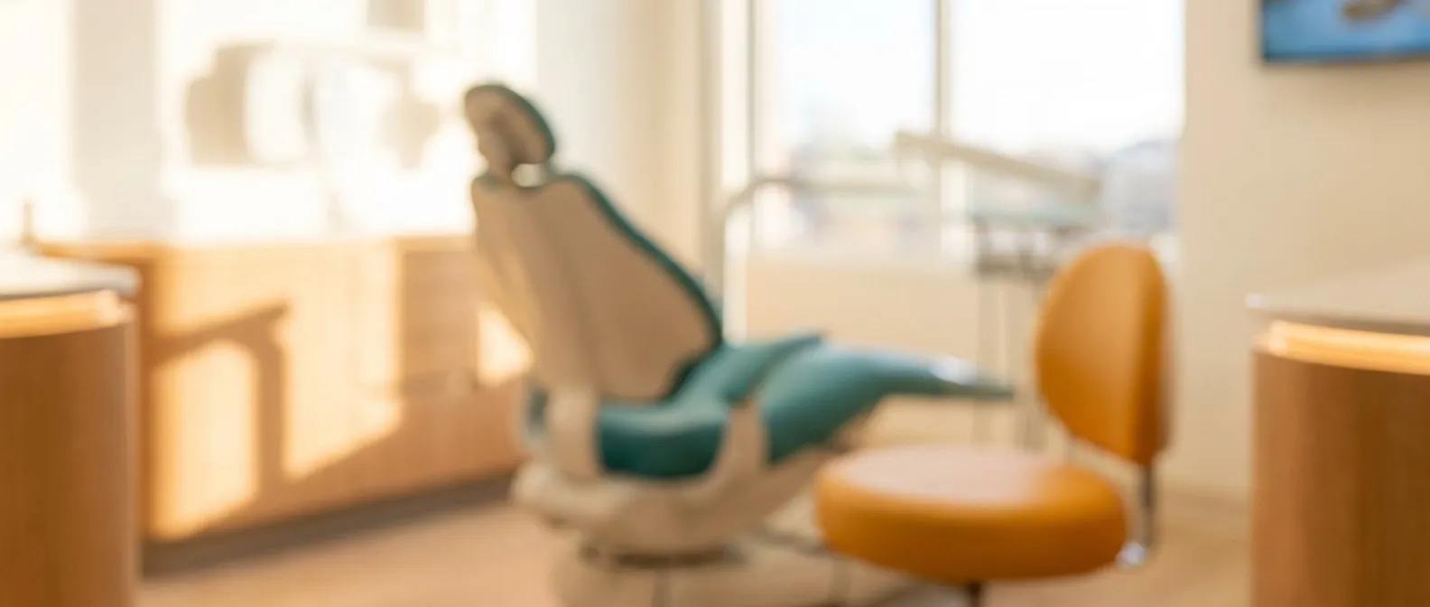 A softly lit treatment room with a teal dental chair and an amber stool, photographed out of focus